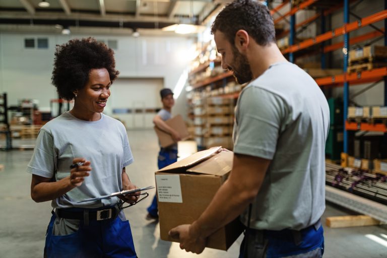 Happy black female worker going through check list and talking to her coworker who is carrying cardboard box in distribution warehouse.